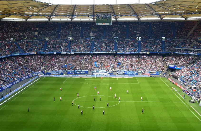 Vista ampla de um estádio de futebol lotado, com torcedores nas arquibancadas e jogadores posicionados no gramado verde para o início de uma partida.