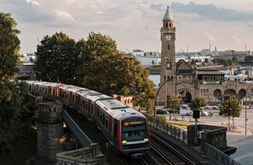 Trem passando pela estação de Landungsbrücken, com a torre do relógio ao fundo e o porto de Hamburgo visível.