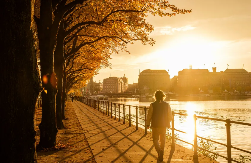 Pessoa caminhando à beira do lago Alster em Hamburgo ao pôr do sol, com árvores de folhas alaranjadas criando uma atmosfera outonal