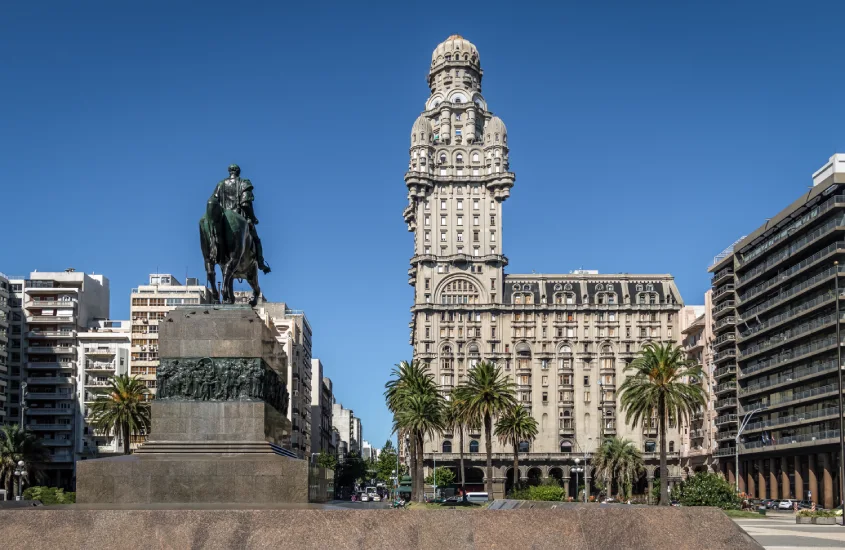 Estátua de José Artigas montado a cavalo na Plaza Independencia, com o icônico Palácio Salvo ao fundo, cercado por edifícios modernos e palmeiras, em Montevidéu