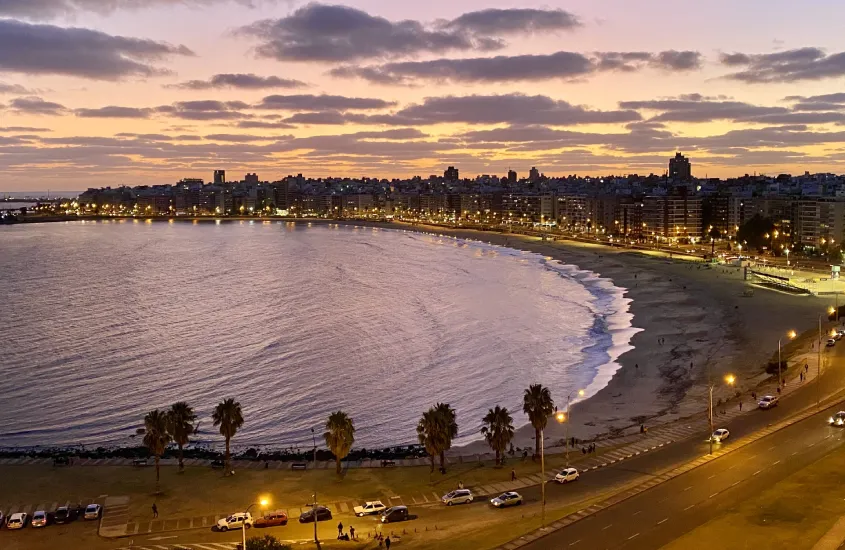 Pôr do sol colorido sobre a Rambla de Montevidéu, com as luzes da cidade e da Playa Pocitos se acendendo, criando um cenário urbano iluminado à beira do Rio da Prata