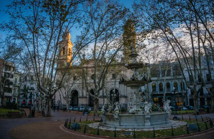 Vista da Plaza Constitución ao entardecer, com árvores sem folhas e a fonte central em destaque, tendo a Catedral Metropolitana e prédios históricos como pano de fundo, no coração da Ciudad Vieja