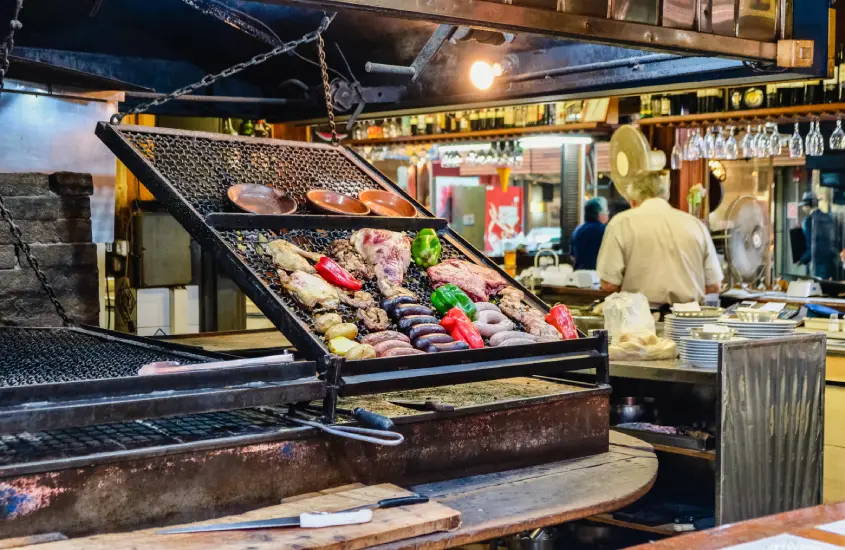 Grelha cheia de cortes de carne e legumes coloridos em uma parrilla tradicional no Mercado del Puerto, um dos locais mais famosos para provar a culinária uruguaia