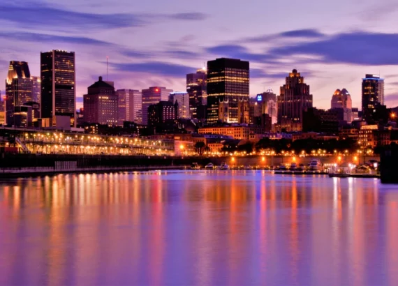 Vista do horizonte de Montreal com os prédios iluminados refletindo nas águas tranquilas do rio, enquanto o céu roxo e azul dá um tom suave ao cenário urbano