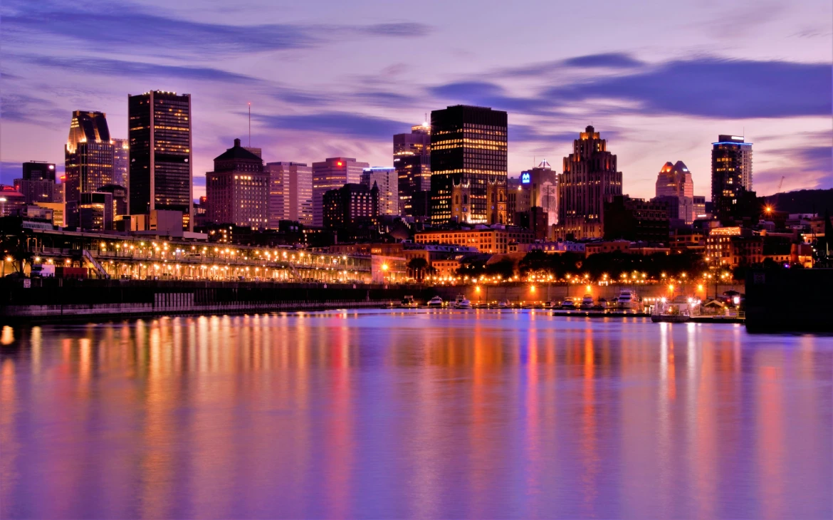 Vista do horizonte de Montreal com os prédios iluminados refletindo nas águas tranquilas do rio, enquanto o céu roxo e azul dá um tom suave ao cenário urbano