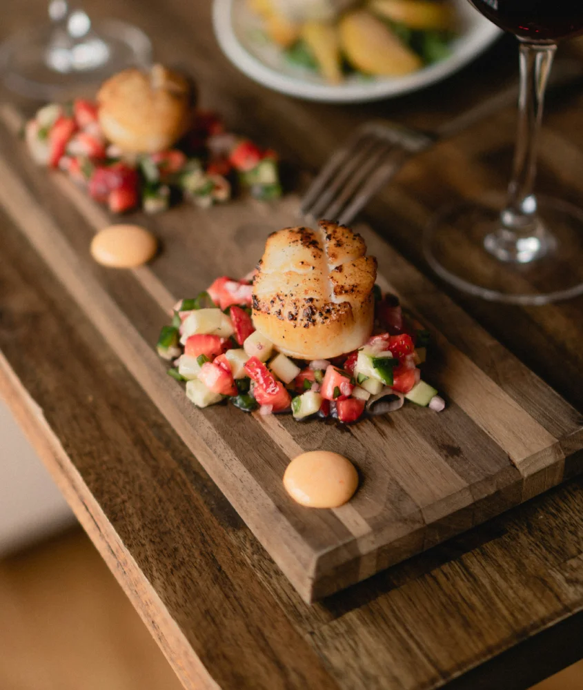 Vieiras grelhadas servidas sobre uma salada fresca de frutas e legumes, acompanhadas por taças de vinho, criando uma experiência gastronômica sofisticada