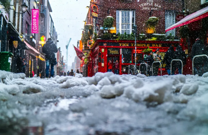 Rua de Temple Bar, em Dublin, coberta de neve, com uma fachada vermelha iluminada ao fundo