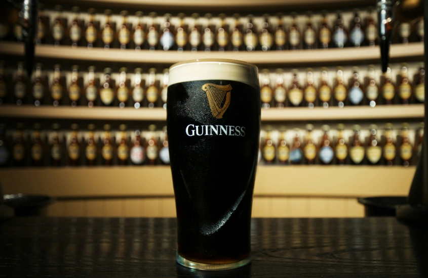 Copo de cerveja Guinness em destaque, com prateleiras de garrafas ao fundo