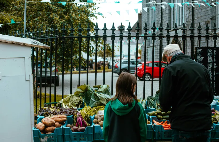 Cena de uma feira ao ar livre com uma jovem e um homem observando uma banca de vegetais frescos, em uma cidade irlandesa