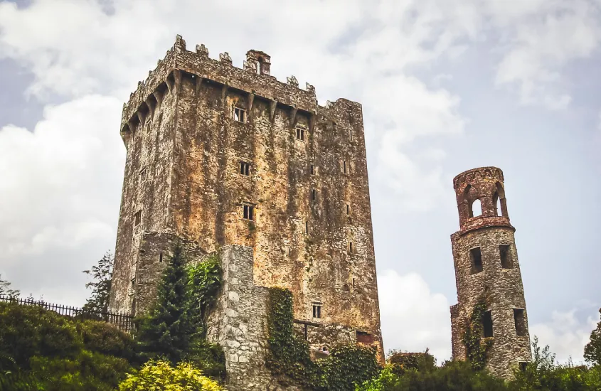 Imponente estrutura do Castelo de Blarney, na Irlanda, cercado por vegetação e com céu nublado ao fundo