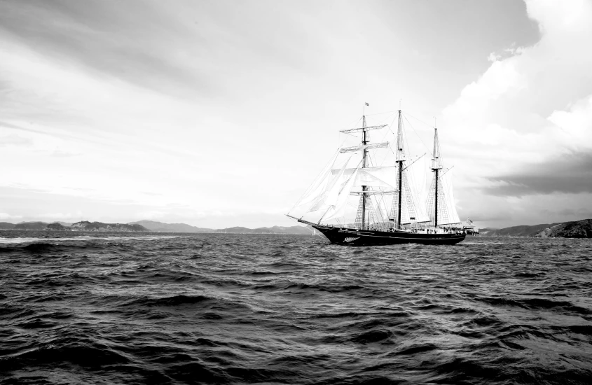 Navio à vela navegando em águas abertas com montanhas ao fundo em um cenário monocromático.