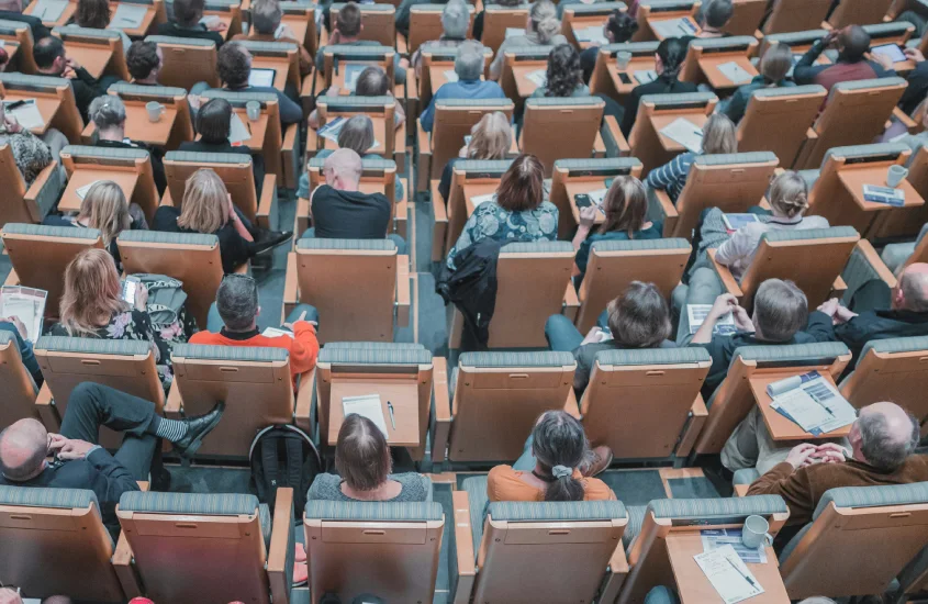 Auditório cheio de pessoas assistindo a uma conferência ou palestra, visto de cima.