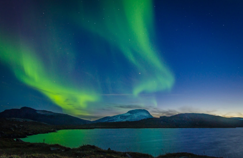 Aurora boreal em tons de verde dançando no céu noturno acima de um lago sueco cercado por montanhas.