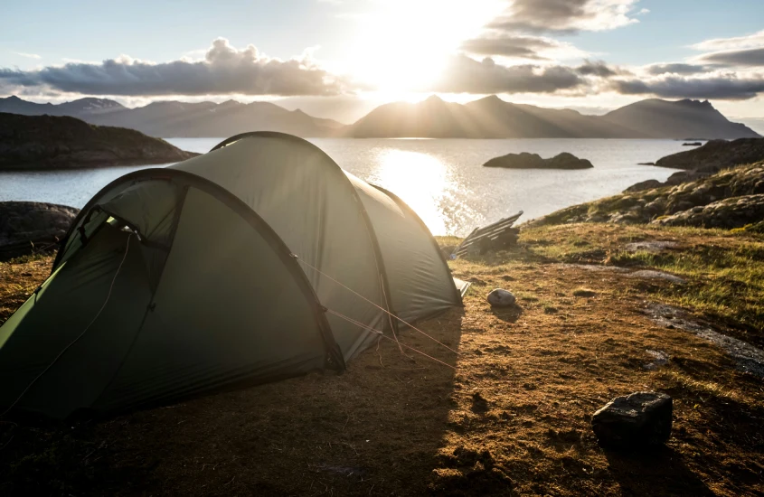 Tenda de acampamento montada à beira de um lago, cercada por montanhas, com o sol brilhando ao amanhecer na Suécia.