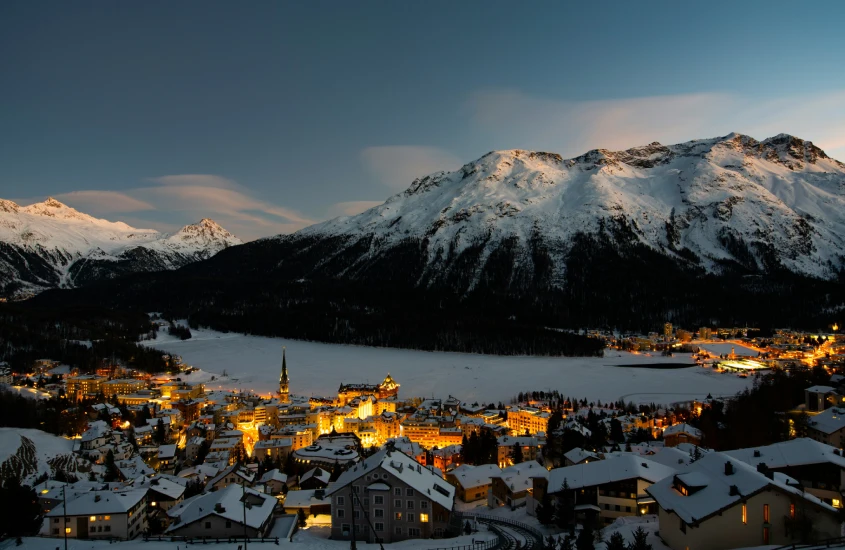 Vista panorâmica noturna de uma cidade cercada por montanhas cobertas de neve iluminadas ao anoitecer