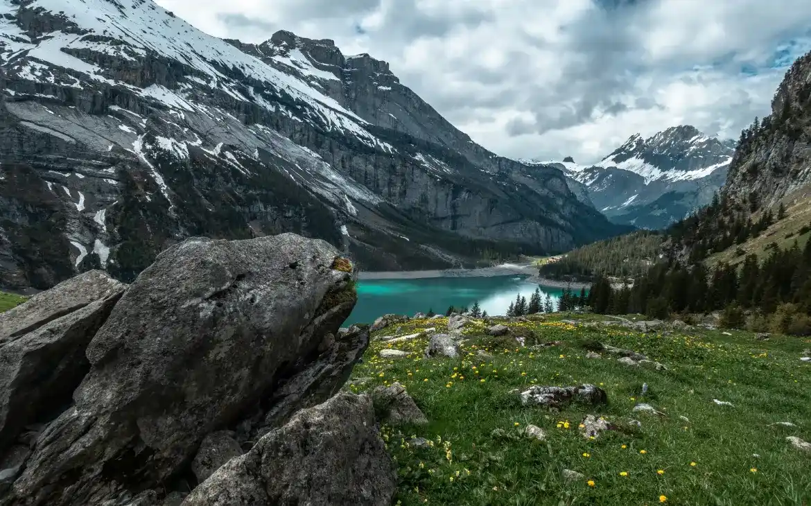 Lago de águas esverdeadas cercado por montanhas com picos nevados e vegetação alpina, com flores amarelas em primeiro plano e grandes rochas no caminho