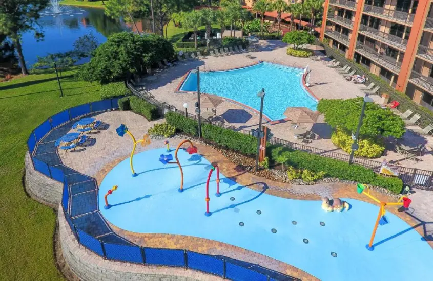 Vista aérea do Rosen Inn Lake Buena Vista com piscina e área infantil de recreação cercada por árvores e cadeiras de sol, com lago ao fundo.