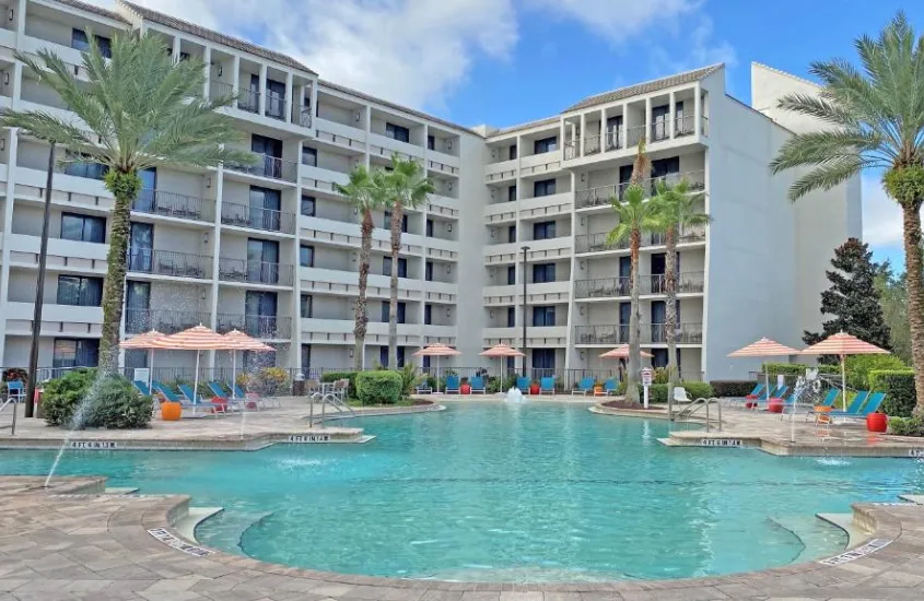 Piscina ampla no Holiday Inn Orlando Disney Springs Area, cercada por guarda-sóis coloridos e cadeiras de sol, com palmeiras ao redor.