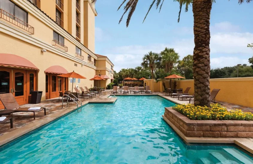 Piscina no Embassy Suites by Hilton Orlando International Drive Convention Center cercada por espreguiçadeiras e prédios ao fundo.