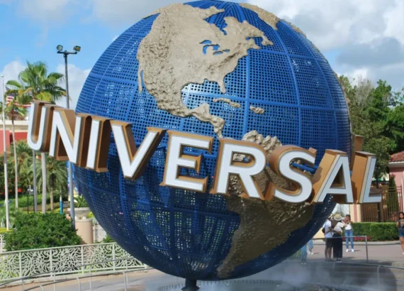 Globo icônico da Universal Studios em destaque na entrada do parque, com detalhes em dourado e azul, cercado por visitantes tirando fotos e bandeiras ao fundo