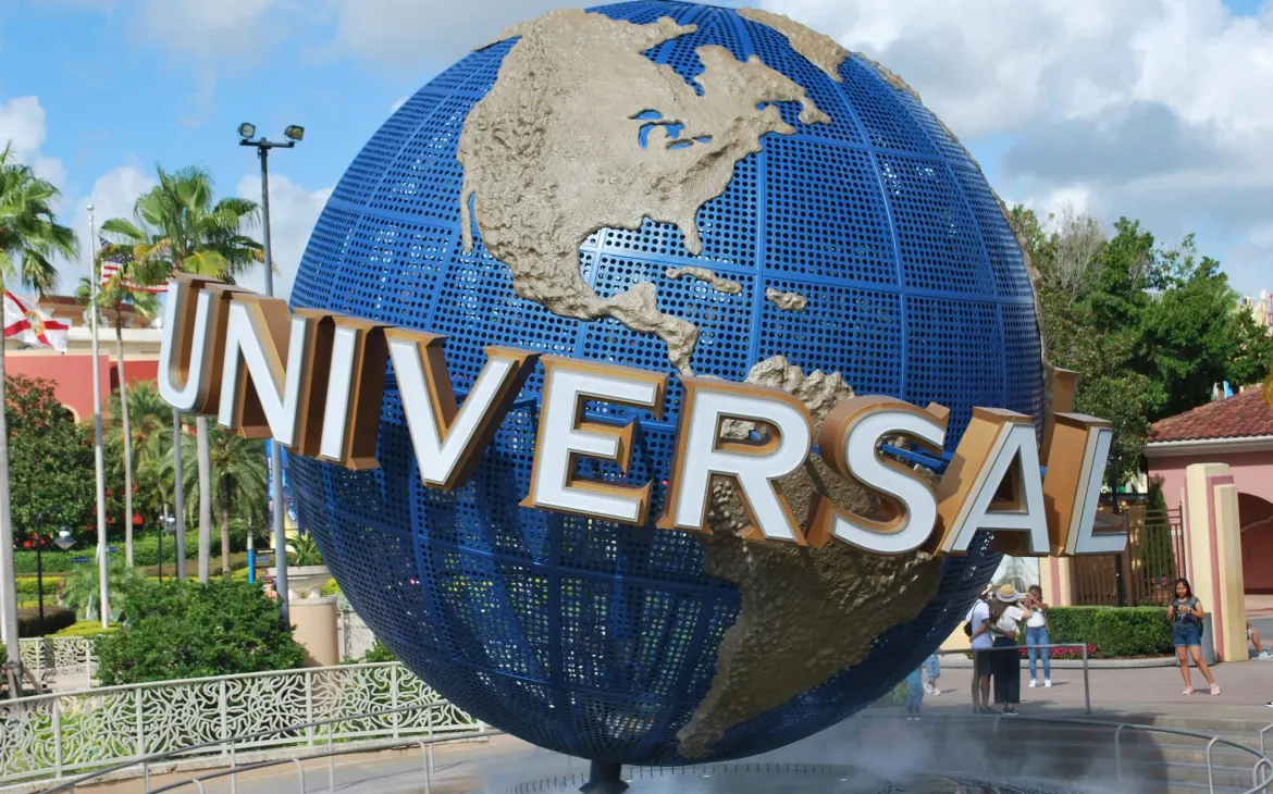 Globo icônico da Universal Studios em destaque na entrada do parque, com detalhes em dourado e azul, cercado por visitantes tirando fotos e bandeiras ao fundo