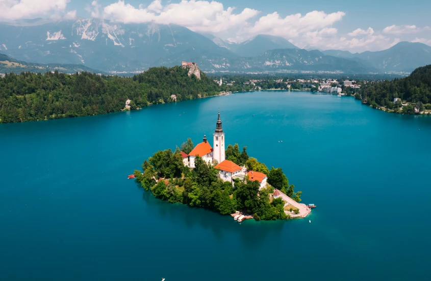 A pequena ilha no centro do Lago Bled, com uma igreja de torre alta cercada por águas azuis cristalinas e montanhas ao fundo