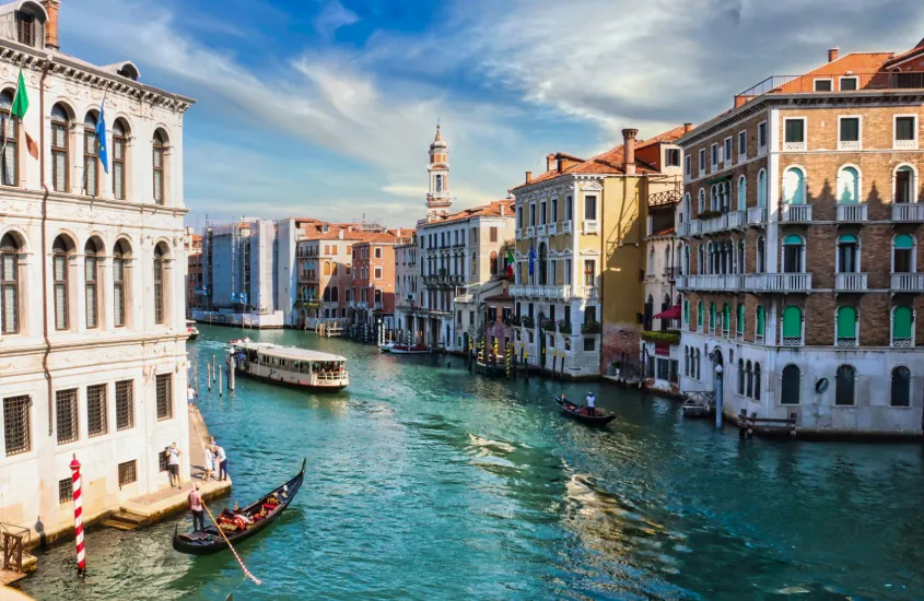 Canal de Veneza com uma gôndola passando entre edifícios históricos, sob um céu azul com nuvens, e um barco de transporte ao fundo