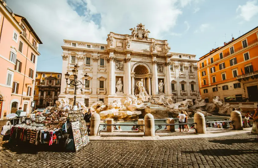 Fonte de Trevi em Roma, com turistas ao redor e uma pequena barraca de souvenirs à frente, em uma manhã ensolarada