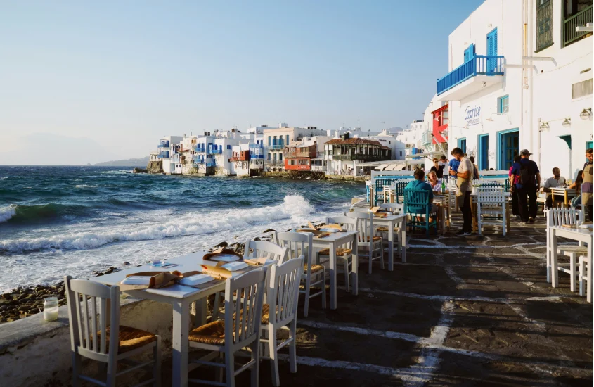 Mesas dispostas à beira-mar em Mykonos, Grécia, com o mar azul e construções brancas típicas da ilha ao fundo