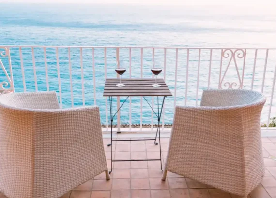 Duas taças de vinho sobre uma mesa em varanda com vista para o mar. Cadeiras de vime branco cercam a mesa.