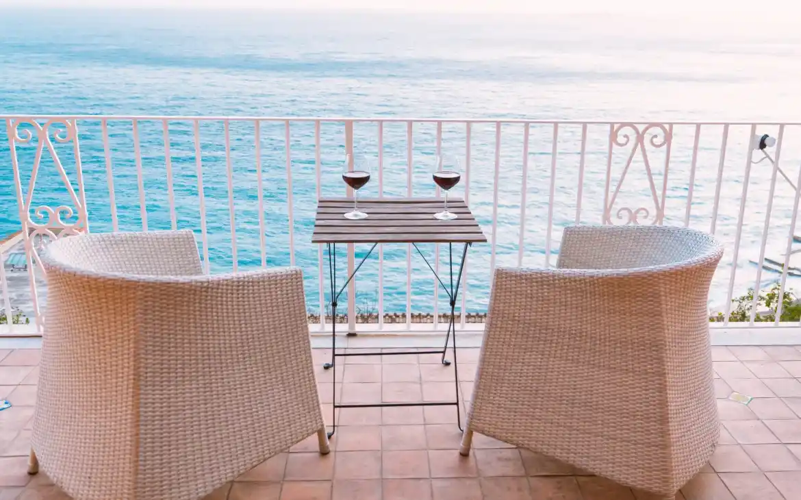 Duas taças de vinho sobre uma mesa em varanda com vista para o mar. Cadeiras de vime branco cercam a mesa.