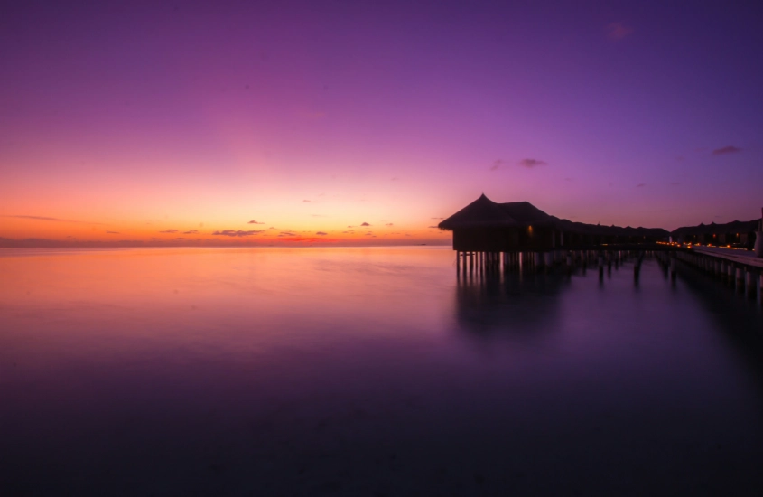 Pôr do sol vibrante em um resort sobre a água, com tons de roxo e laranja refletindo na superfície calma do mar