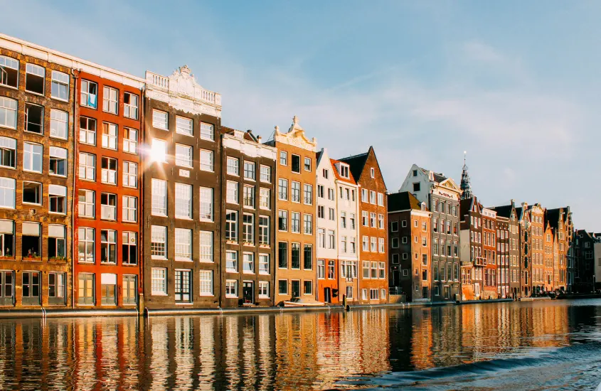 Edifícios coloridos à beira de um canal em Amsterdã, refletindo na água em um dia ensolarado, criando um cenário romântico