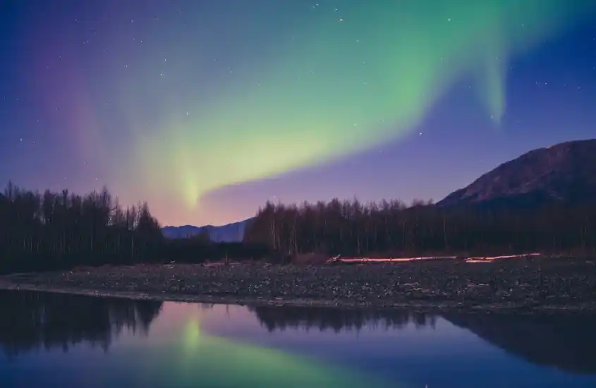 Uma linda aurora boreal ilumina o céu noturno com tons de verde e roxo, refletindo em um tranquilo rio cercado por árvores