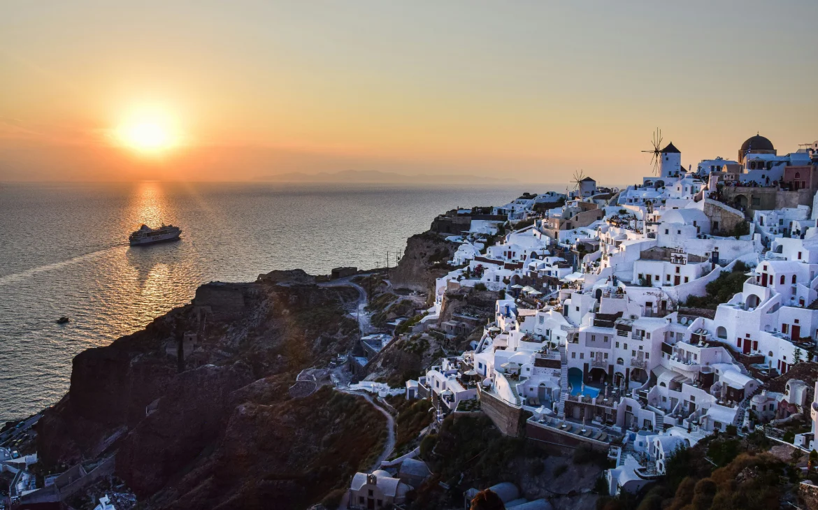 Santorini ao pôr do sol, com casas brancas em estilo grego e um barco navegando nas águas calmas do mar Egeu