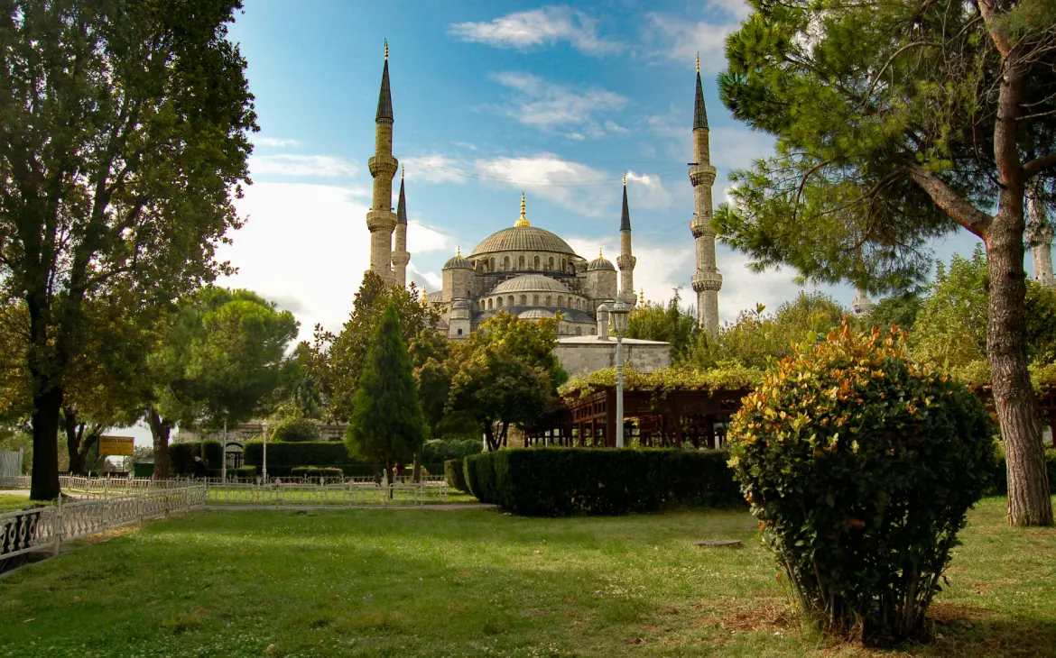 Vista da Mesquita Azul em Istambul, com seus minaretes e cúpula dourada emoldurados por árvores e jardins no primeiro plano. Céu claro ao fundo complementa a paisagem