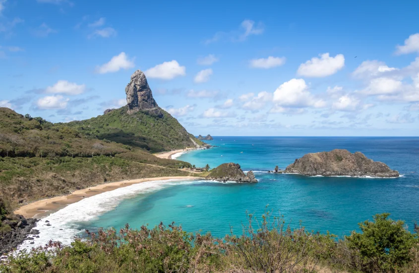 Vista de praia em Fernando de Noronha com mar azul-turquesa e a icônica formação rochosa do Morro do Pico ao fundo, sob um céu azul com nuvens esparsas.