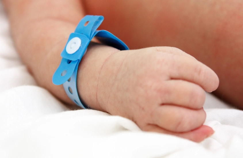 Mão de um bebê com pulseira de identificação hospitalar azul, representando a delicadeza e o começo da vida.