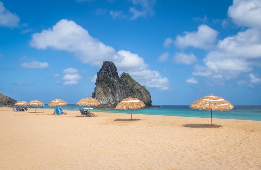 Praia de areia dourada com guarda-sóis de palha e vista para a icônica formação rochosa, destacando um dia ensolarado em Noronha.