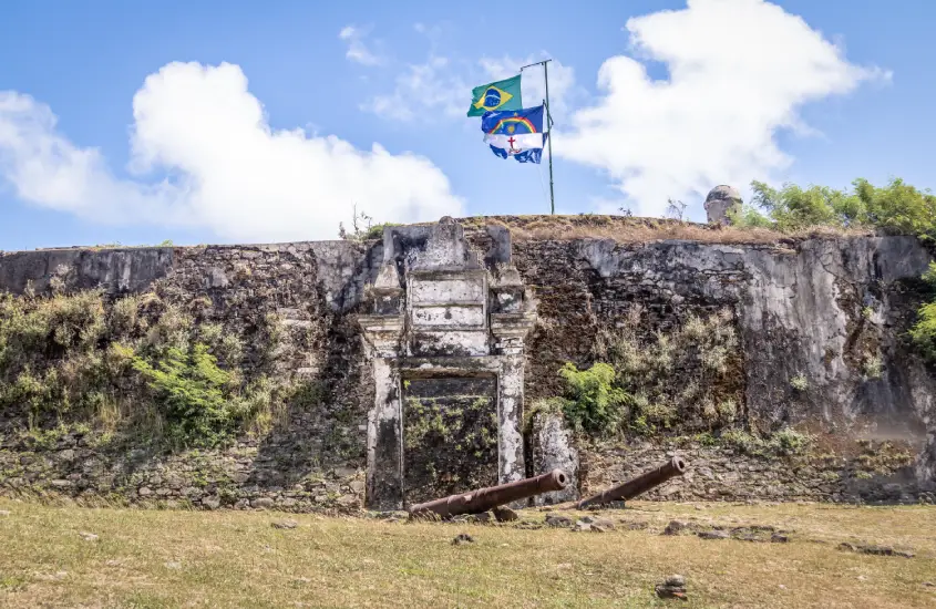 Bandeiras do Brasil e de Pernambuco tremulando sobre uma antiga fortificação, realçando a história e o orgulho local.