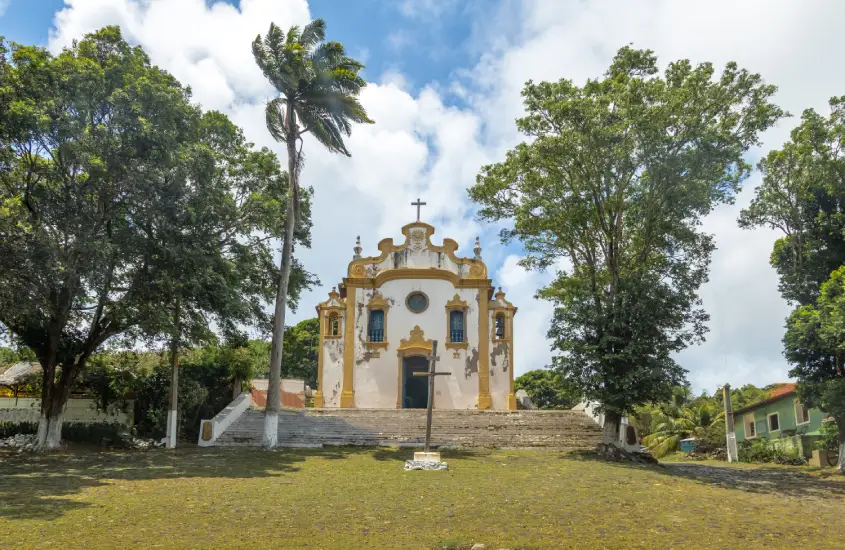 Igreja colonial no alto de uma colina, rodeada por árvores e palmeiras, com uma cruz em destaque no pátio frontal.