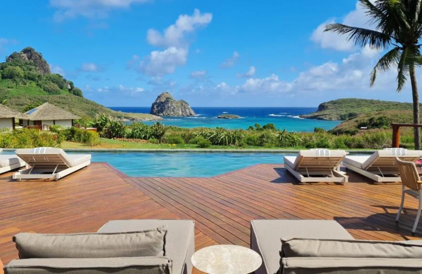 Piscina de borda infinita com espreguiçadeiras de madeira, oferecendo vista para o mar e a vegetação de Noronha.