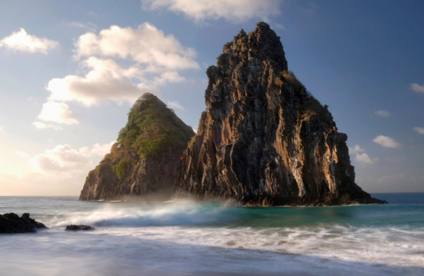 Imponente formação rochosa dos Dois Irmãos emergindo do mar em Fernando de Noronha, com ondas suaves batendo na base, ao amanhecer.