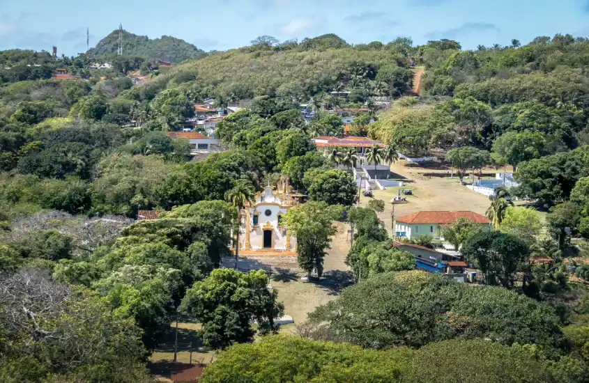 Vista aérea da Igreja de Nossa Senhora dos Remédios, em Fernando de Noronha, cercada pela vegetação densa da ilha e com o povoado ao fundo.