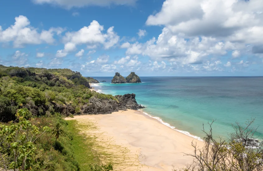 Praia deserta com areia clara, vegetação verde ao redor e o mar em tons de azul e verde, com as ilhas Dois Irmãos ao fundo, compondo um cenário paradisíaco e tranquilo.