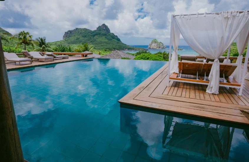 Piscina de borda infinita com vista para montanhas e mar em Fernando de Noronha, com espreguiçadeiras e um gazebo decorativo.