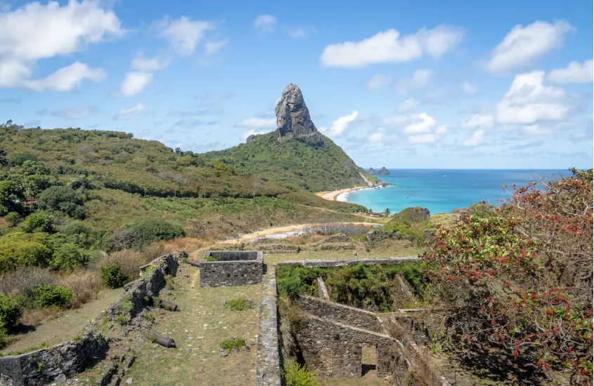 Ruínas históricas em Fernando de Noronha, com vista para o Morro do Pico e o mar ao fundo.