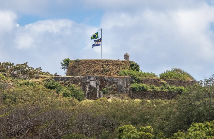 Forte de Fernando de Noronha com bandeira do Brasil e de Pernambuco tremulando ao vento, cercado por vegetação.