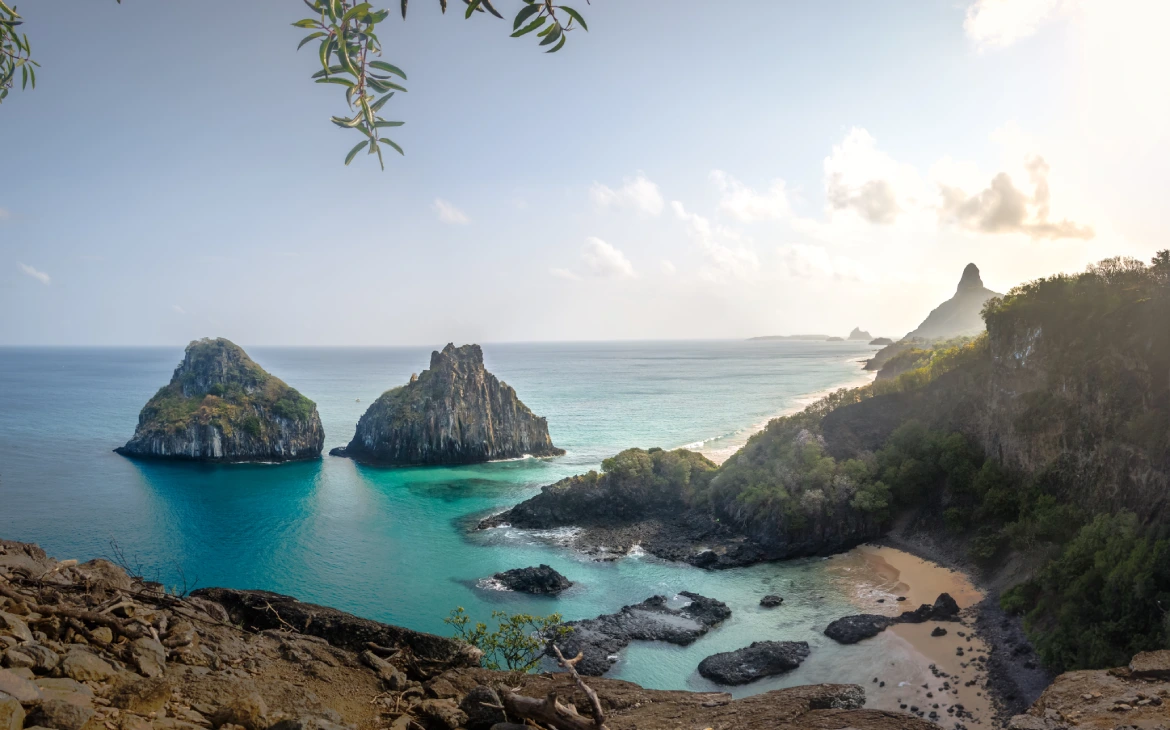 Os morros Dois Irmãos em Fernando de Noronha, cercados pelo mar azul e vegetação ao redor, com o Pico visível ao fundo.