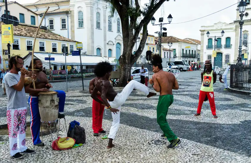 Capoeiristas demonstrando movimentos no Pelourinho, com músicos tocando berimbau e tambor, em uma praça histórica de Salvador.